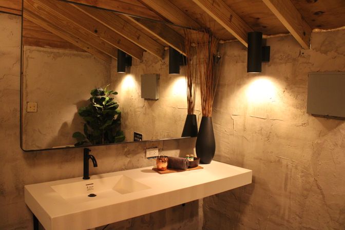 Cozy modern-rustic bathroom with a long white rectangular sink and matte black faucet, large mirror, textured plaster walls, exposed wood-beam ceiling, warm wall sconces, tall black vases with dried reeds and a potted plant.
