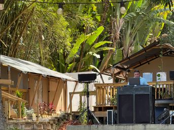 DJ at an outdoor tropical glamping site, laptop and speakers on a wooden deck surrounded by banana palms and canvas tents