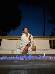 Laughing woman wrapped in a white blanket on a backyard patio sofa, toasting a marshmallow over a blue-flamed outdoor fire pit at twilight.