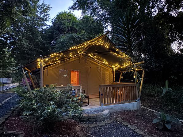 Cozy canvas glamping tent with wooden porch and warm string lights at dusk in a lush tropical forest retreat.