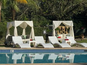 Inviting sunlit poolside with white curved lounge chairs and two draped cabanas framed by tropical palms and green hedges, reflected in calm blue water.