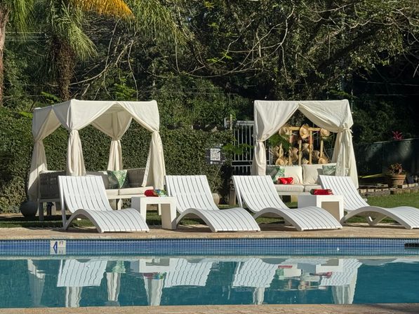 Inviting sunlit poolside with white curved lounge chairs and two draped cabanas framed by tropical palms and green hedges, reflected in calm blue water.