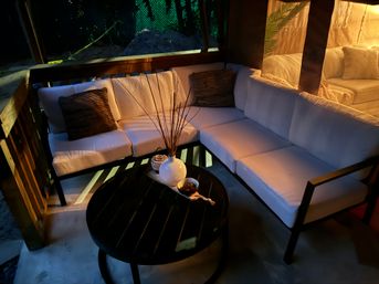 Cozy backyard patio lounge with L-shaped white cushioned sofa on a wooden deck, round black coffee table holding a white vase with reeds and a small bowl, warm evening ambient lighting and throw pillows.