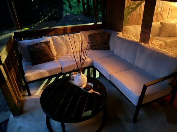 Cozy backyard patio lounge with L-shaped white cushioned sofa on a wooden deck, round black coffee table holding a white vase with reeds and a small bowl, warm evening ambient lighting and throw pillows.