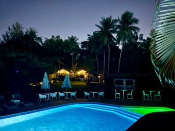 Nighttime tropical resort scene with a glowing blue-green pool, silhouetted palm trees, string-lit bungalows and patio tables under a starry sky.