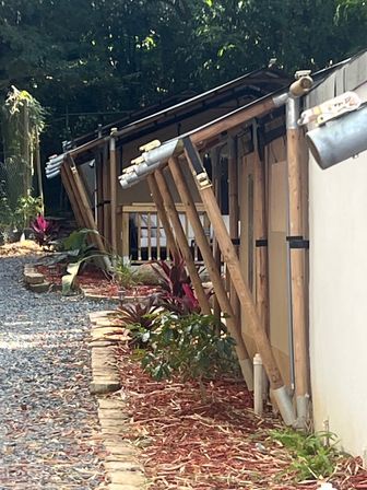 Sun-dappled gravel path beside a light-colored building with angled wooden supports propping a sloped metal-gutter roof, stone edging and red-mulch beds planted with tropical foliage under a shady tree canopy.