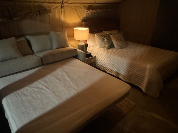Cozy dimly lit room with a made bed and a pulled-out sofa bed, warm bedside lamp, neutral quilted bedding and decorative pillows.