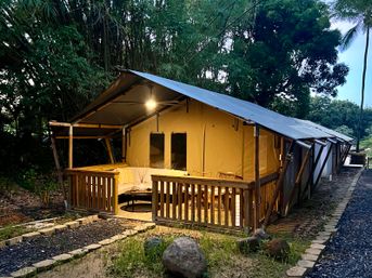 Cozy safari-style glamping tent with wooden porch and warm lit lounge at dusk, nestled in tropical bamboo and palm trees