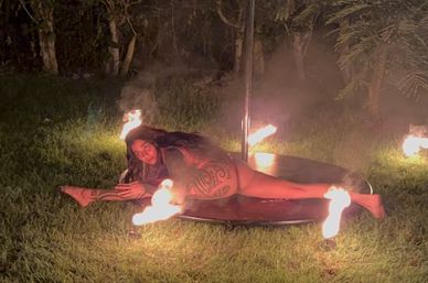 Tattooed pole dancer in a bikini stretched across a circular pole platform at night, surrounded by small ground flames on grassy outdoor fire performance