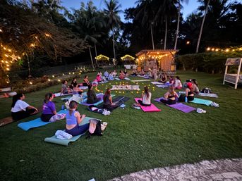 Relaxing outdoor evening yoga and meditation circle on a grassy lawn at sunset, participants on colorful mats around candles and string lights under tall palm trees at a tropical retreat.