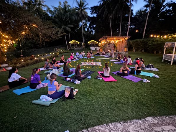 Relaxing outdoor evening yoga and meditation circle on a grassy lawn at sunset, participants on colorful mats around candles and string lights under tall palm trees at a tropical retreat.