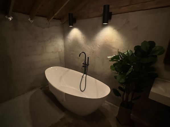 Moody rustic bathroom with a white freestanding tub, black freestanding faucet, two black wall spotlights, textured concrete walls, exposed wood ceiling beams, and a potted fiddle-leaf fig plant.