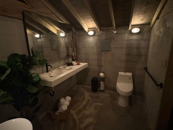 Moody industrial-style concrete bathroom with exposed wooden ceiling beams, wall-mounted rectangular sink with black faucet, white toilet, caged wall lights, potted plant and wicker basket of rolled towels.
