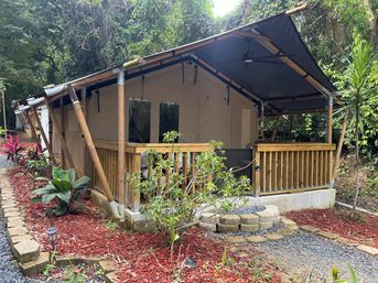 Bamboo-framed glamping tent with wooden porch and ceiling fan nestled in lush tropical forest, gravel path and red-mulch landscaping.