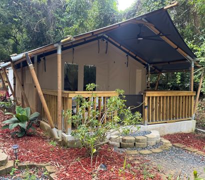 Bamboo-framed glamping tent with wooden porch and ceiling fan nestled in lush tropical forest, gravel path and red-mulch landscaping.