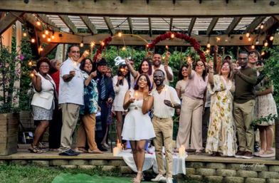 Couple in white and beige pose in front of family and friends on a decorated backyard pergola at an evening garden celebration, guests holding sparklers beneath string lights and a floral arch.