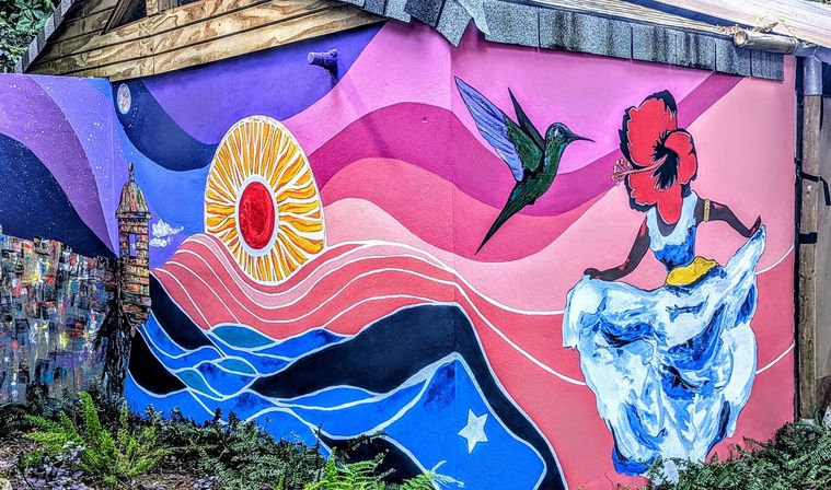 Vibrant outdoor mural on a garden wall showing a radiant sun over pink-and-blue waves, a hummingbird near a hibiscus-headed dancer in a flowing white-blue dress.
