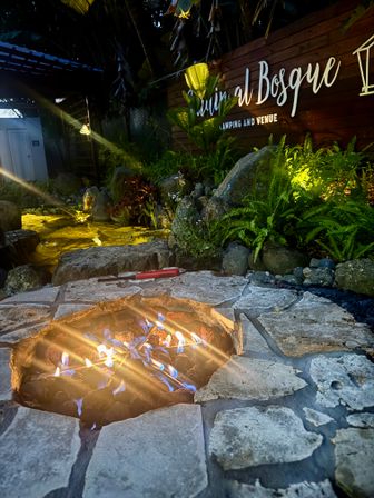 Stone fire pit with blue-yellow flames in a cozy tropical garden patio at night, glowing pond, rocks and lush ferns lit by warm landscape lighting