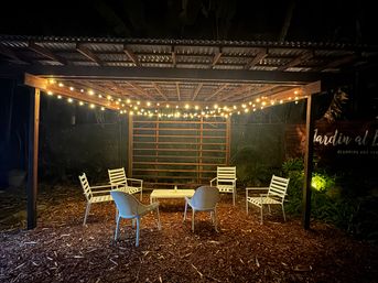 Cozy outdoor pergola glowing with warm string lights, white patio chairs and a low table arranged on wood-chip ground in a nighttime garden seating nook.