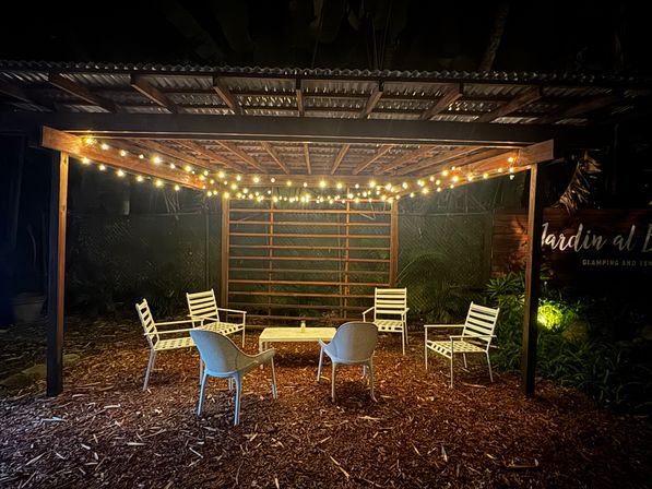 Cozy outdoor pergola glowing with warm string lights, white patio chairs and a low table arranged on wood-chip ground in a nighttime garden seating nook.