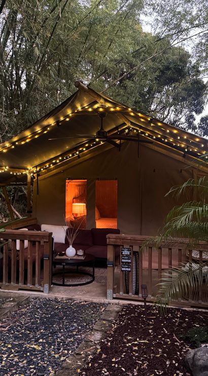Cozy glamping canvas tent patio aglow with string lights and a warm lamp inside, nestled in a tropical garden with palm trees and a stone path.