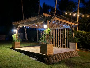 Cozy wooden backyard pergola on a raised stone-edged deck with planter boxes and blue string lights at night, framed by palm trees and a grassy lawn.