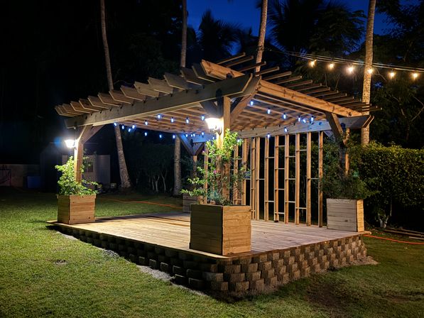 Cozy wooden backyard pergola on a raised stone-edged deck with planter boxes and blue string lights at night, framed by palm trees and a grassy lawn.