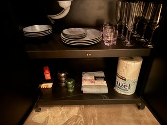 Under-sink kitchen cabinet with stacked plates, wine glasses and tumblers, a paper towel roll, folded dish towel and small utility items beneath visible sink plumbing.