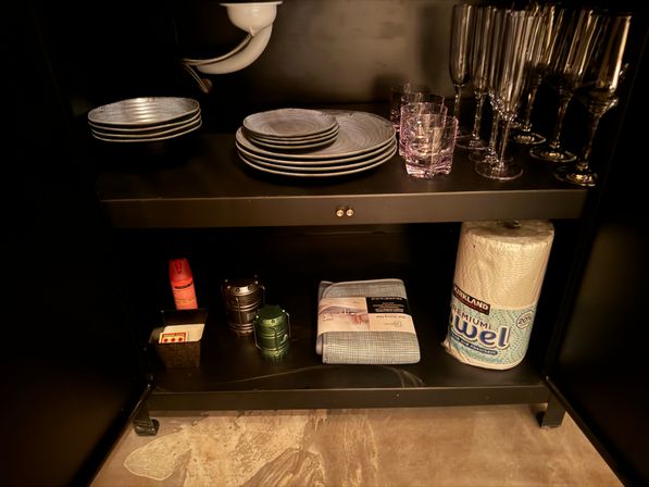Under-sink kitchen cabinet with stacked plates, wine glasses and tumblers, a paper towel roll, folded dish towel and small utility items beneath visible sink plumbing.