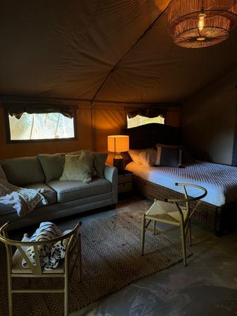 Cozy glamping tent interior with canvas walls, wicker bed and quilt, sofa with textured pillows, warm bedside lamp, woven rug and chairs — rustic, inviting campsite bedroom lounge.