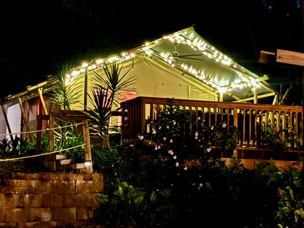 Nighttime glamping tent on a raised wooden deck decorated with warm string lights, tropical plants and flowering shrubs in a garden setting