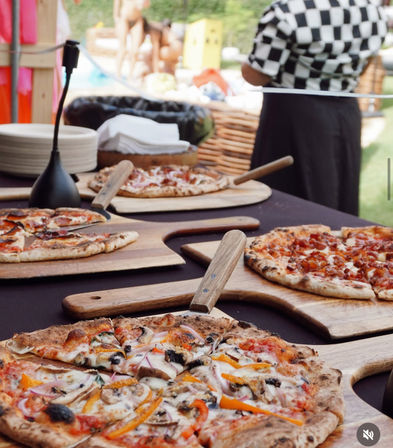 Assorted wood-fired pizzas with vegetable and meat toppings displayed on rustic wooden pizza peels at an outdoor pizza buffet, blurred server in checkered shirt in background