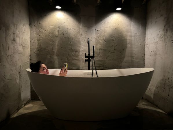 Person relaxing in a freestanding white bathtub in a minimalist concrete-walled bathroom under two dim spotlights, holding a smartphone — modern urban spa vibe.