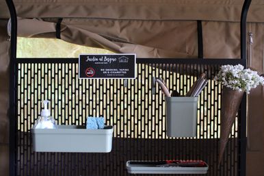 Perforated black metal organizer on a tan outdoor canopy wall holding sage-green trays with a soap dispenser and sponge, a utensil cup and tools, a cone of white flowers, and a small no-smoking sign.