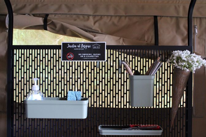 Perforated black metal organizer on a tan outdoor canopy wall holding sage-green trays with a soap dispenser and sponge, a utensil cup and tools, a cone of white flowers, and a small no-smoking sign.