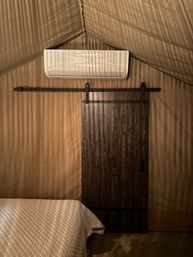 Cozy glamping tent interior with quilted bed, dark wood sliding barn door on a black track, wall-mounted mini-split AC, and warm striped lighting on tan canvas walls and ceiling.
