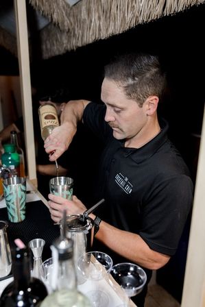 Bartender pouring liquor into a cocktail shaker at a tiki-style bar, mixing tools, jigger and coupe glasses visible on the counter
