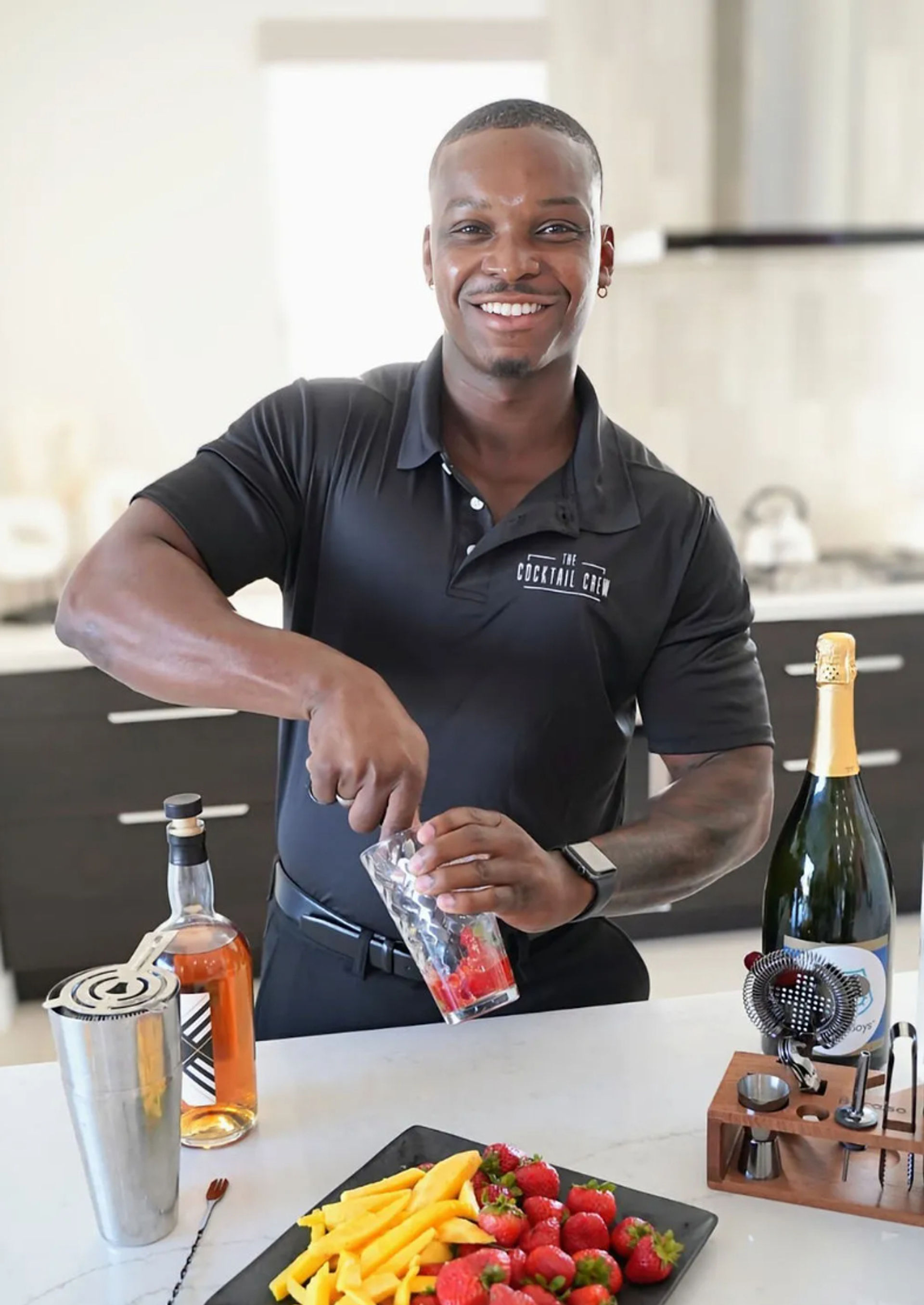 Smiling bartender muddling strawberries in a glass at a modern kitchen island with bottles, cocktail tools and a plate of fresh strawberries and mango slices