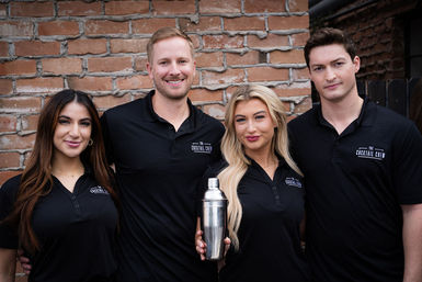 Four smiling bartenders in matching black polos pose against a brick wall with one holding a stainless steel cocktail shaker, urban team photo