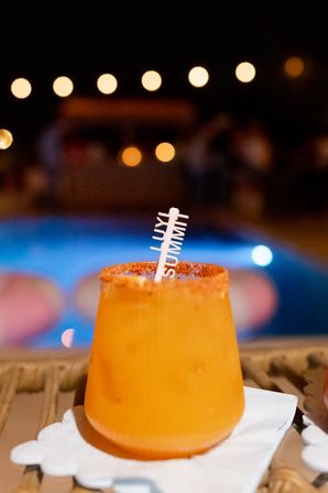 Poolside zesty orange cocktail with chili-rim and decorative stirrer on a napkin, blurred lit swimming pool and string‑light bokeh at night