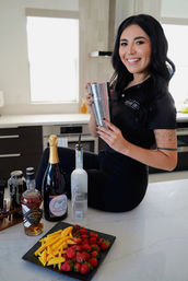 Smiling woman on a modern kitchen island holding a stainless steel cocktail shaker, with bottles of spirits and champagne and a black plate of sliced mango and fresh strawberries on the countertop.