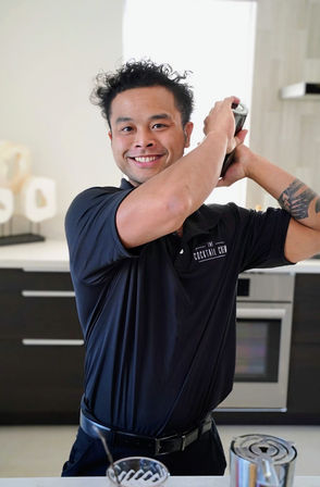 Smiling bartender energetically shaking a cocktail shaker in a bright modern kitchen, wearing a black polo and showing a geometric forearm tattoo.