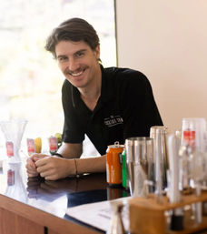 Smiling bartender with a mustache leaning on an indoor bar counter, surrounded by cocktail shakers, mixers, glasses and cans