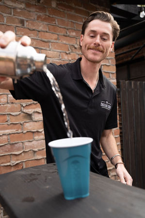 Smiling bartender in black polo pours a cocktail from a metal shaker into a blue plastic cup on a rustic brick outdoor patio.