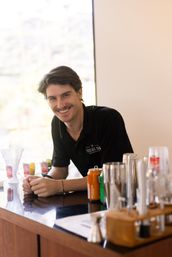 Smiling bartender leaning on a glossy wooden bar counter with cocktail shakers, canned mixers and glasses in a sunlit modern cocktail bar.