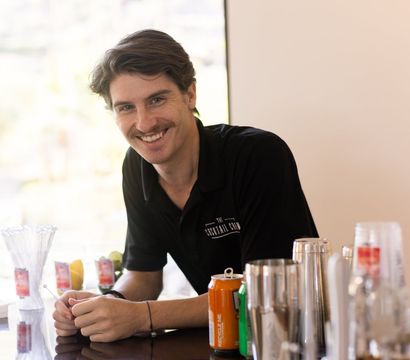 Smiling bartender leaning on a sunlit bar counter with cocktail shakers, soda cans, glassware, and fresh fruit garnishes at a cocktail station.