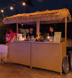 Nighttime outdoor tiki-style bar on a stone patio with a thatched roof, string lights overhead and two people behind a rattan counter serving cocktails — cozy backyard party setup