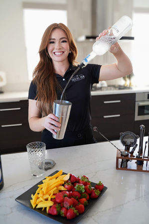 Smiling bartender pouring clear liquor into a metal cocktail shaker in a modern home kitchen bar, with a plate of strawberries and mango slices on a marble countertop.