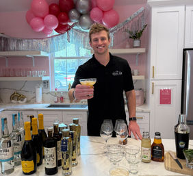 Smiling bartender in a black polo offers a yellow cocktail across a marble countertop in a modern white kitchen decorated with pink and silver balloons, surrounded by bottles, glassware and bar tools for a celebration.