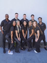 Ten-person smiling staff team in matching black polo shirts and black pants posing outdoors against a light gray wall — upbeat office team portrait.